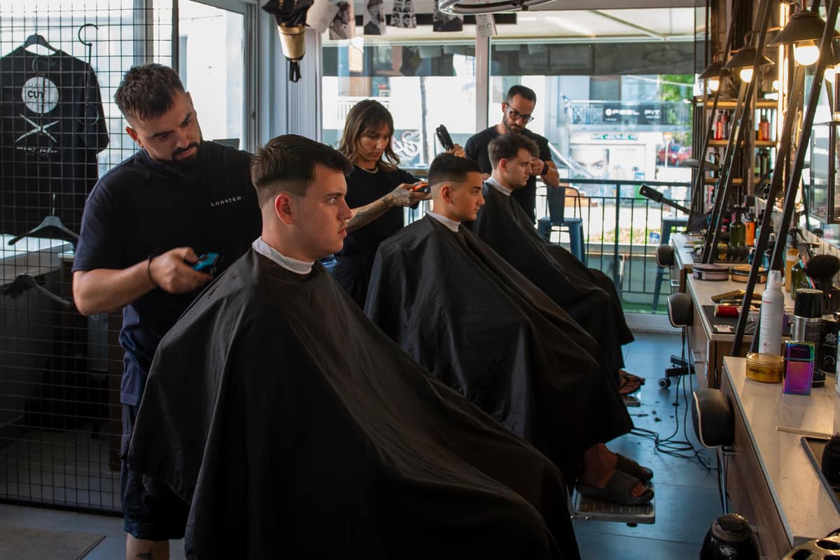 Μέσα στο Haircut Fresh: τρεις barber κουρεύουν στις καρέκλες μπροστά στα μεγάλα παράθυρα, κάτω από το μαύρο πλέγμα στο ταβάνι